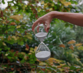 A glass hourglass held by a man's hand from above. Green vegetal background. The sand is white....