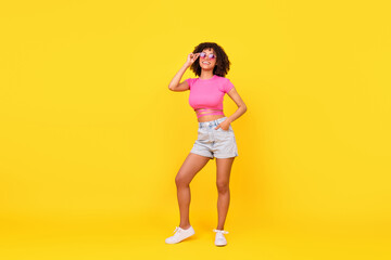 Full length photo of shiny dreamy girl dressed pink t-shirt dark glasses looking empty space isolated yellow color background
