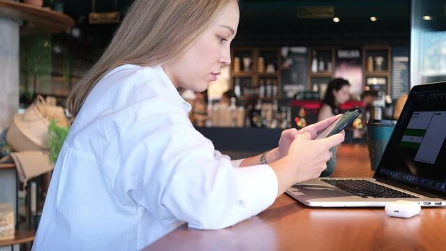 Young woman in white shirt sitting in cafe with phone in hands typing working on laptop freelance businesswoman chatting message