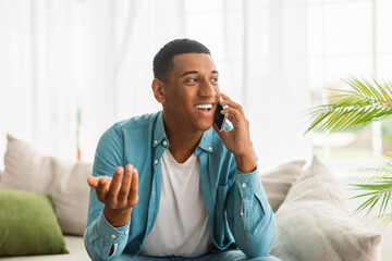 Smiling millennial african american male gesturing hand and calling by smartphone in white living room interior