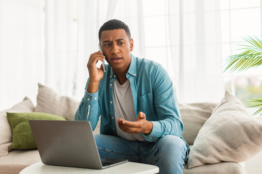 Unhappy serious busy young african american male work with computer, calls by smartphone