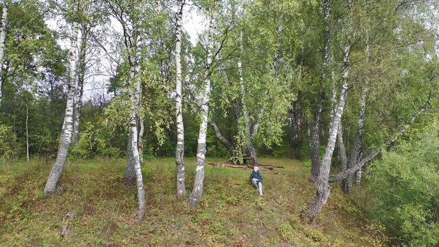 Young blondie woman in a green jacket is sitting on the ground. Beautiful autumn forest with birches. Drone view of landscape with colorful trees and golden leaves. Drone shooting footage