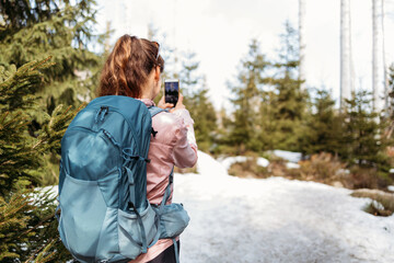 A girl tourist with a backpack on her back takes a photo on a mobile phone while walking in the forest, doing hiking. Using gadgets while hiking.
