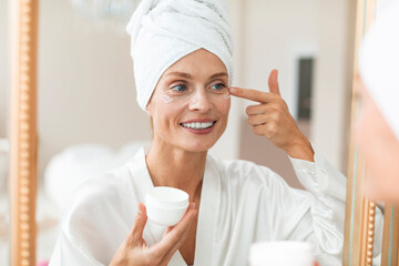 Beautiful middle aged woman in silky bathrobe applying moisturizing eye cream, enjoying face care routine - obrazy, fototapety, plakaty