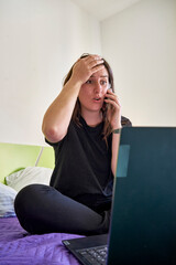 Vertical shot of the worried white woman talking with a phone and sitting on the bed
