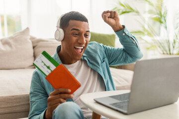 Glad excited young black male in wireless headphones holding passport and air tickets, doing victory