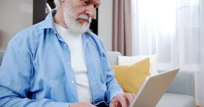 Close-up beautiful elderly man holding using laptop and smiling. Handsome senior sitting on sofa in living room typing on keyboard. Busy old male with gray hair and beard. Gadget concept.