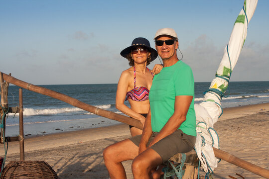 Mature Couple At The Beach In Combuco, Brazil, Sitting On A Jangada A Fishing Boat Typical In Brazil