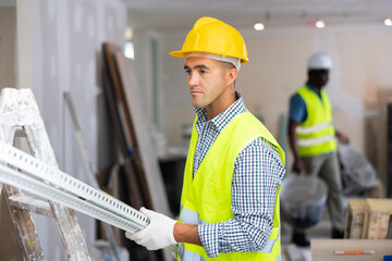 Repairman wearing protective hardhat and yellow vest carrying construction detail while working in...