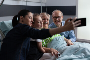 Adult woman taking self portrait with smartphone accompanied of elderly patient, relatives in geriatric hospital room. Family selfie with sick grandmother at clinic visiting area.