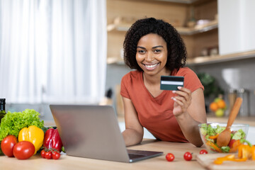 Smiling young black woman in red t-shirt with computer show credit card at table with vegetables