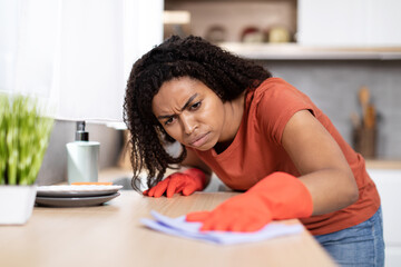 Dissatisfied sad young black lady in red t-shirt, rubber gloves wipes dirt from wooden table in kitchen interior
