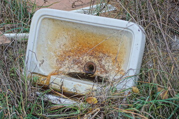 garbage from one white old ceramic washstand sink in brown dirt stands in green and gray dry grass...