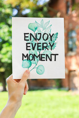 Woman holding card with phrase Enjoy Every Moment outdoors, closeup