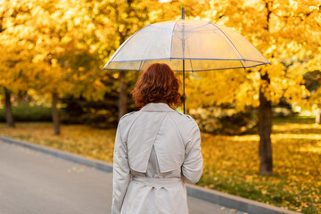 Millennial european red-haired woman in raincoat with umbrella walks in rain, enjoy bad weather alone