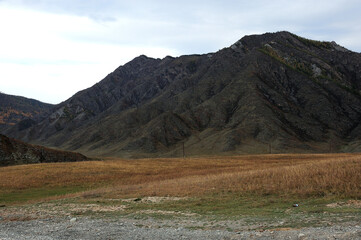 A flat rocky clearing with dried and yellowed grass at the foot of a high mountain.