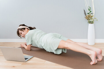 家でパソコンを見ながら腕立て伏せ・オンライントレーニングするパジャマ姿のアジア人女性
