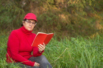beautiful mature woman lies on a green meadow in the forest and reads a book