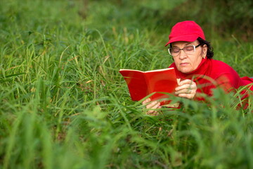 beautiful mature woman lies on a green meadow in the forest and reads a book