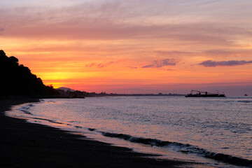 View of glowing fiery sunset of pink, orange and blue overlooking peaceful ocean and silhouette...