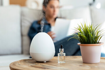 Shot of essential oil aroma diffuser humidifier diffusing water articles in the air while woman reading a book sitting on coach. - obrazy, fototapety, plakaty