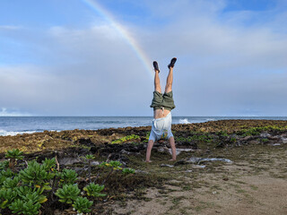 Man does Handstand on coral rocks on the beach with rainbow in the sky