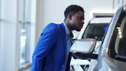 African American man client examines modern car and thinks about purchasing in automobile salon. Black customer tries to search nice vehicle
