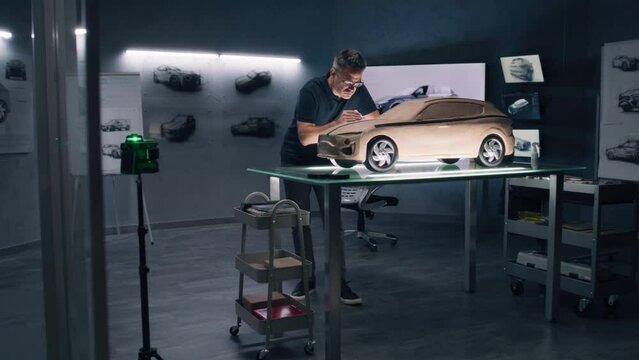 Two male automotive designers creating model of electric car in modern car design studio. One working in 3D modeling computer program, other sculpting with plasticine clay and rake.