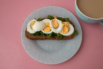 On blue plate there is grain bread with greens and an egg and a cup of coffee on a pink background the concept of a proper nutrition