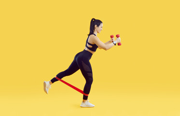 Fototapeta na wymiar Sportswoman working out with elastics ribbon and with dumbbells on vivid yellow background. Girl in sportswear trains with dumbbells in her hands and with elastic band on legs. Full length. Side view.
