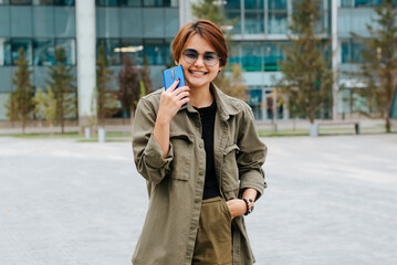 Happy laughing young asian woman holding mobile phone and looking at camera outdoors. Stylish cheerful woman with short haircut with positive emotions on background of modern architecture