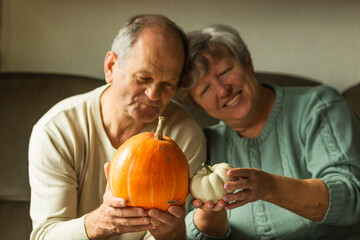 Smiling senior couple holding two pumpkins side by side. Happy elderly family preparing decor to Halloween holiday.