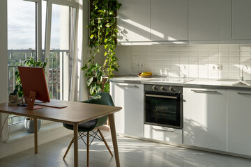 Workplace with monitor for remote employee working from home or doing freelance orders from own apartment. Minimalistic computer desk and office chair in sunny kitchen with houseplant and large window