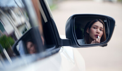 Young woman applying lipstick looking at reflection in car mirror.