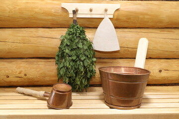 A copper sauna accessories and dry birch broom are on a wooden bench and on a hanger on a log wall in a timber bath.  