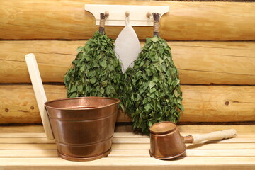 A copper sauna accessories and dry birch brooms are on a wooden bench and on a hanger on a log wall in a timber bath.  