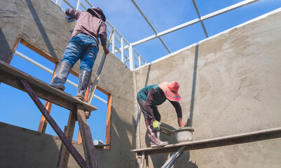 Low angle view of 2 Asian builder workers on wooden scaffolding are plastering concrete wall room...