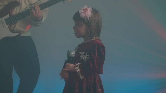 Little girl on stage in vintage dress, she sings into microphone and dances, her father plays an electric guitar. Color music is shining and smoke is billowing. Family time, creativity and hobbies