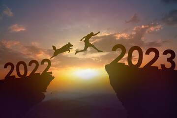 Silhouette dog and young man jumping from 2022 to 2023 at cliff on sunset. New year and success...