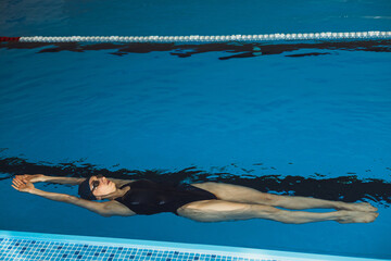 Full length of athletic professional female swimmer showing backstroke while swimming in water of indoor pool during training 