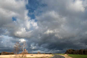 Dramatic sky with dark rain clouds over asphalt country road in autumn landscape