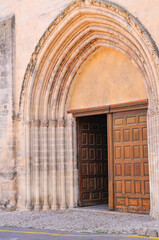 front view, medium distance of, the entrance to a  1500's century cathedral, with carved, limestone walls and two, enormous, hand carved, wood doors, one open to interior