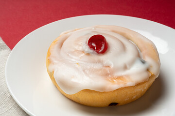 Close-up of a freshly made iced  bun with a glazed cherry on top served on a plate.