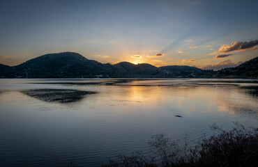 The sun rising the hills over Geni Bay, Lefkada island, Greece with the calm sea water reflection the sunlight and clouds.