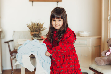 A charming little girl in a dress made of natural material rides a rocking horse in her room....