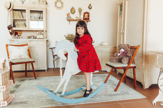 A charming little girl in a dress made of natural material rides a rocking horse in her room. antique nursery interior