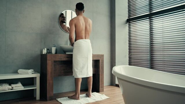 Full length back view naked muscular Indian Latina bearded man guy in bathroom with towel on hips looking at mirror reflection touching beard grooming after shaving check acne face skin pampering