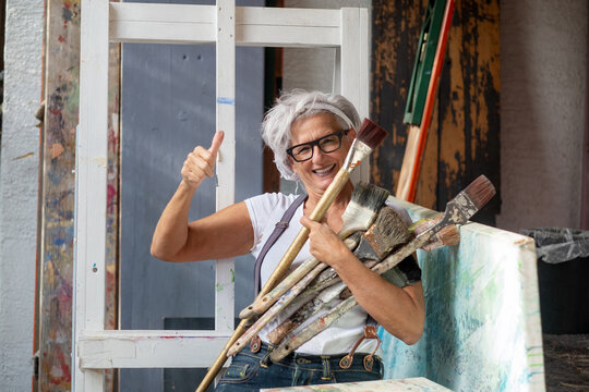 older gray haired mature laughing happy artist woman with glasses and big brushes gives thumbs up