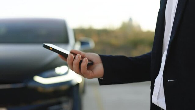Unrecognizable businessman using smartphone app for remote controlling of electric car, driver distantly approaching car, smart car. Sensor scanning road ahead for vehicles, danger, speed limits