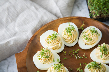 Kalte gefüllte russische Eier mit Frischkäse, Kresse und Kräuter farciert auf Holz Eierteller, Leinen Tuch und Marmor Hintergrund hell zu Ostern, Büffet, Snack oder Vorspeise